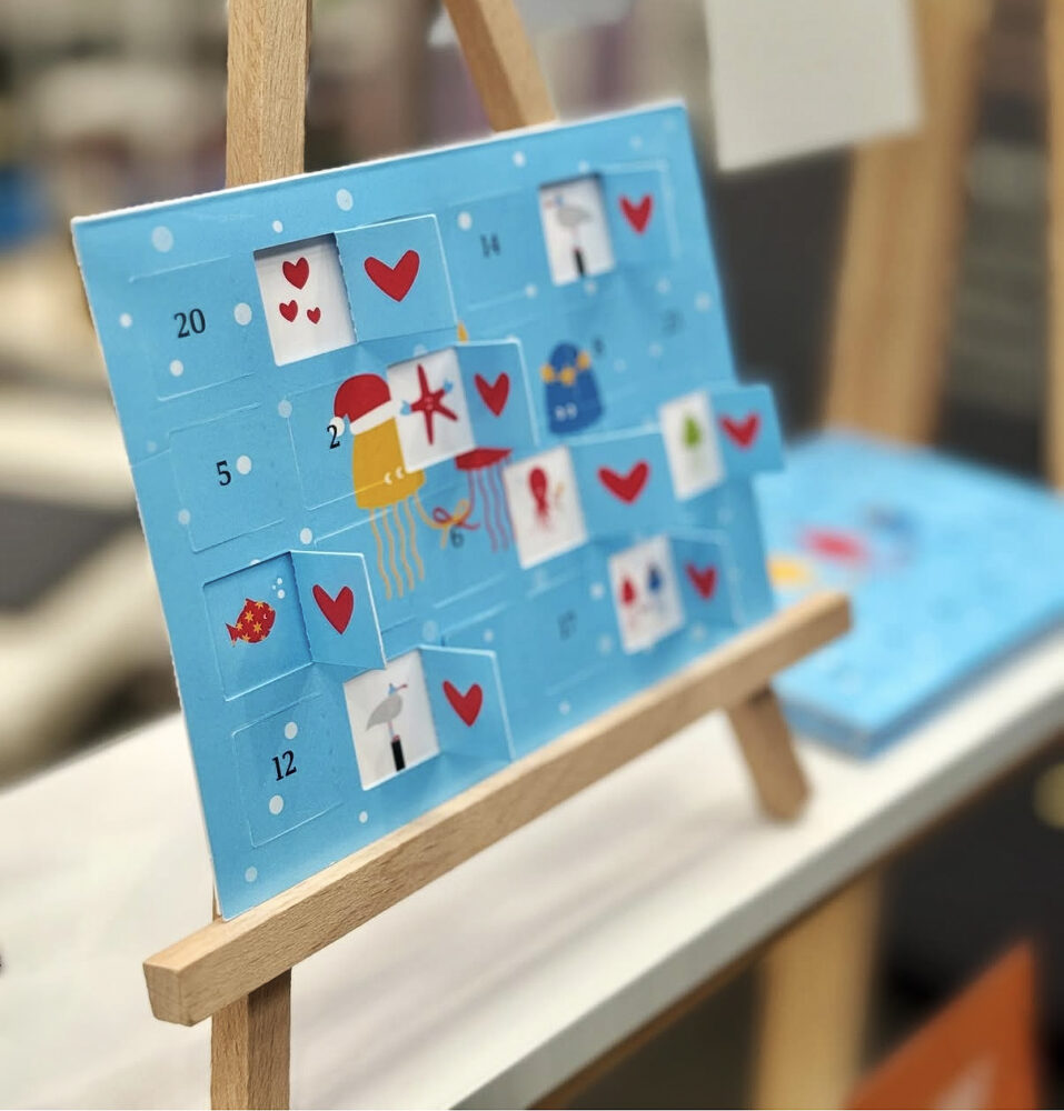 Blauer Adventskalender mit 24 Türchen, winterlichen Motiven und roten Herzen auf einer kleinen Holzstaffelei. Einige Türchen sind bereits geöffnet.