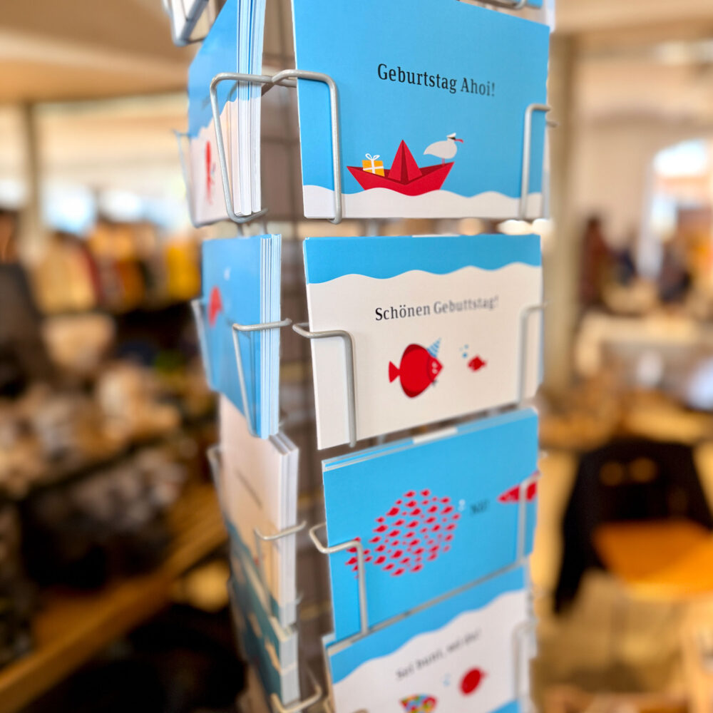 **Alt-Text:**
Drehständer mit mehreren Postkarten in einem Geschäft. Sichtbar sind verschiedene Karten mit blauem Hintergrund und maritimen Motiven, darunter ein rotes Papierboot mit dem Text Geburtstag Ahoi sowie weitere Karten mit Fischen und kurzen Texten.
